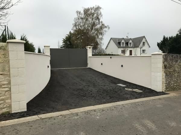 Allée privée fermée par un portail, aux murs blancs et à l'asphalte foncé, menant à une maison en arrière-plan.