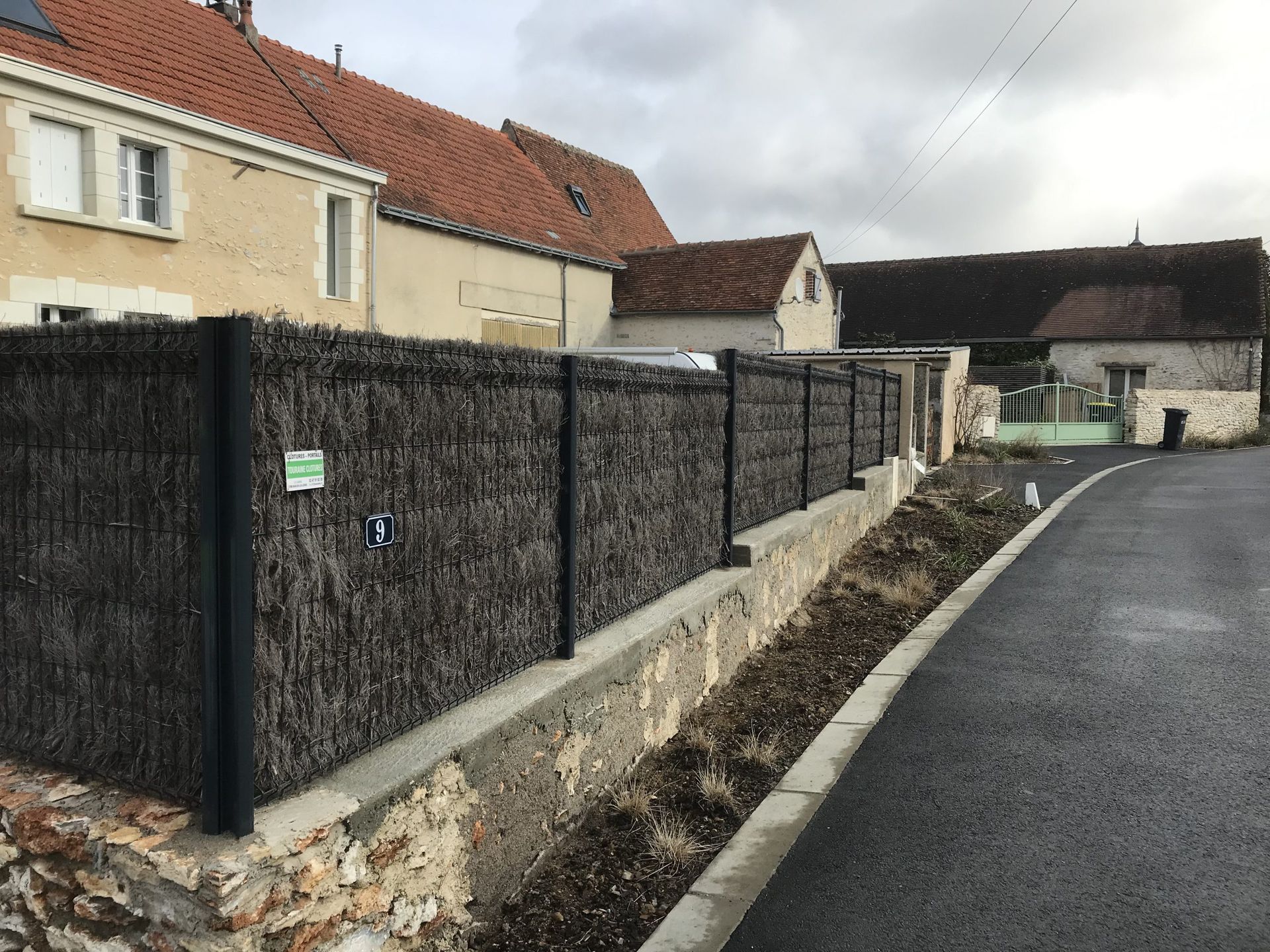 Rue résidentielle bordée d'un mur de pierre et d'une clôture noire, longeant une route pavée sous un ciel nuageux.