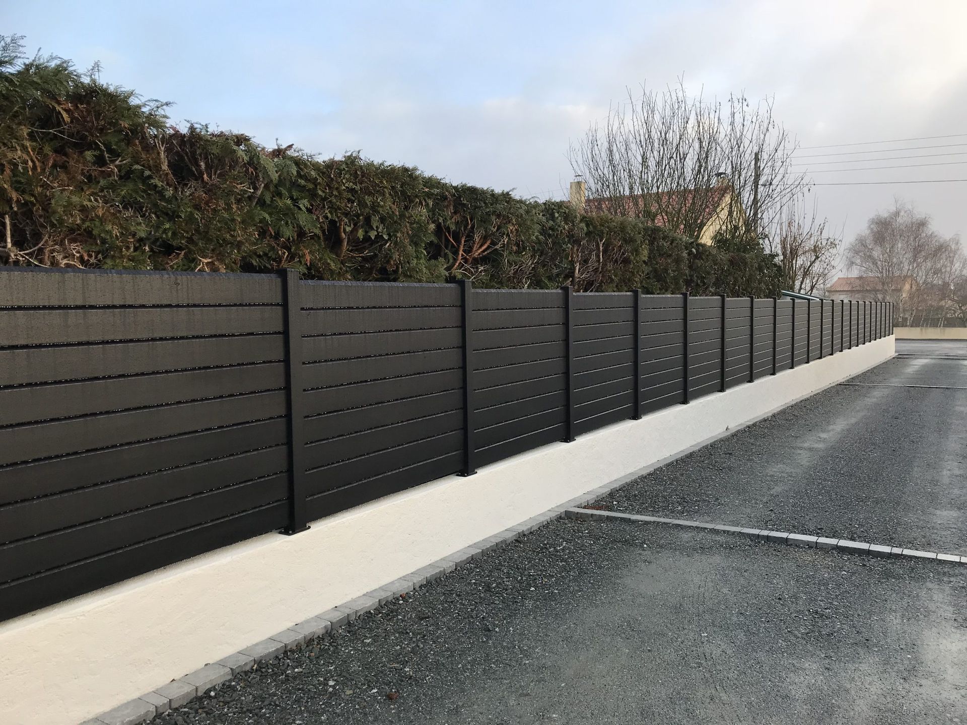Clôture moderne à claire-voie noire le long d'un chemin de gravier, bordée d'un trottoir et de haies blanches.