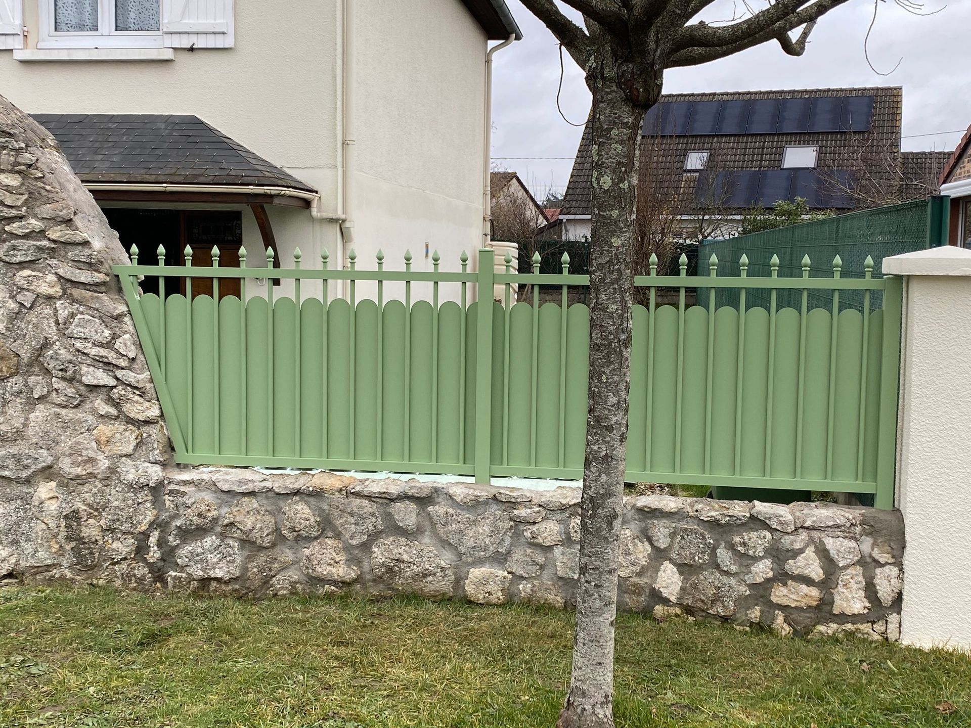 Clôture vert clair avec socle en pierre à côté d'une maison et d'un arbre dans une cour gazonnée