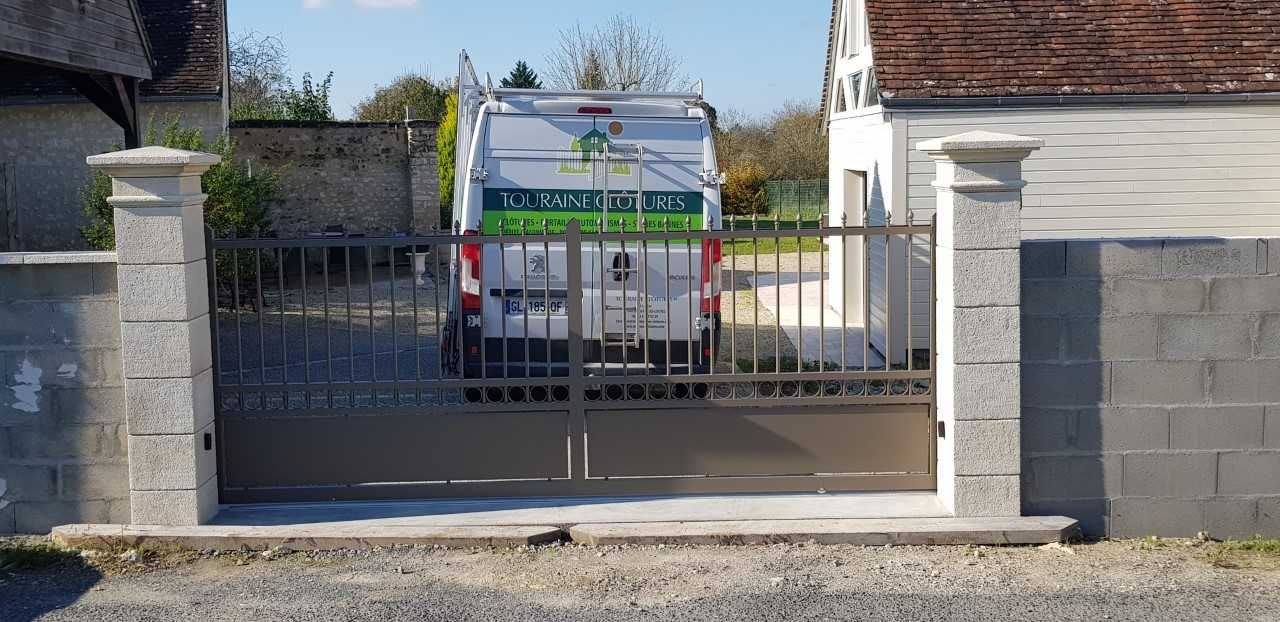 Camion stationné derrière un portail métallique entre des piliers en pierre dans une allée résidentielle.