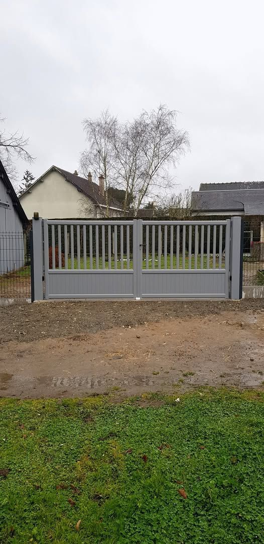 Portail d'allée en métal gris devant une maison, avec une cour en gravier et de l'herbe verte au premier plan.