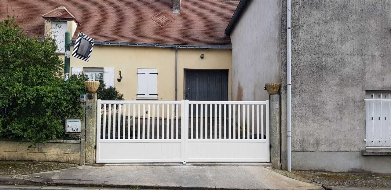 Portail blanc donnant sur une petite maison couleur crème au toit de tuiles rouges et bordée d'un mur gris.