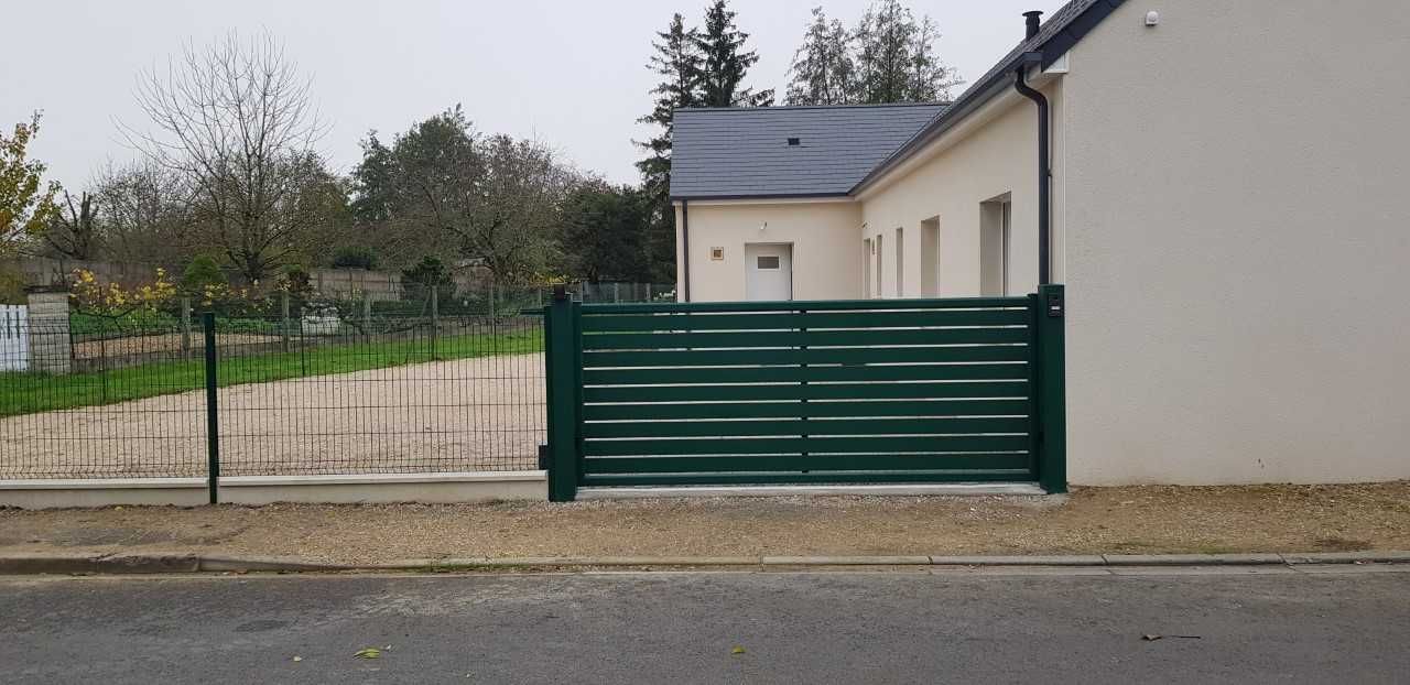Un portail en métal vert se dresse devant une maison beige, au bord d'une route goudronnée, avec une allée de gravier et des arbres à l'arrière.