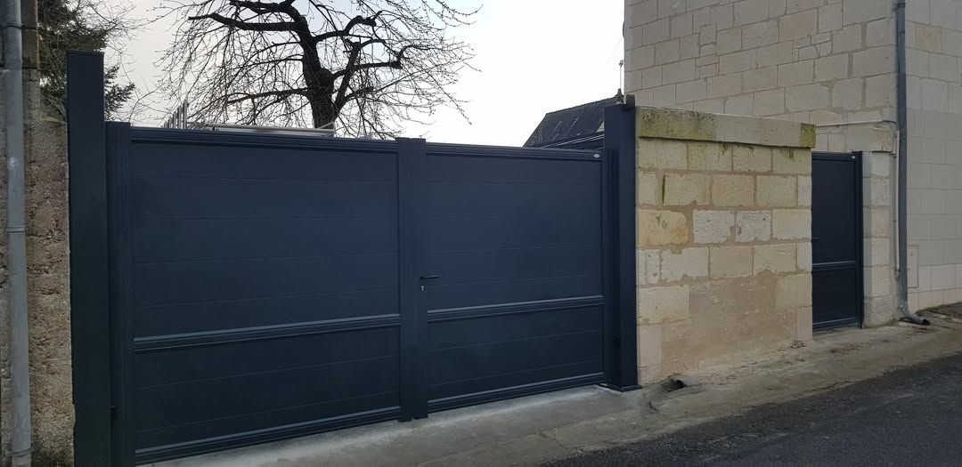 Portail en métal sombre jouxtant un mur de pierre beige dans une rue calme, avec des arbres dénudés en arrière-plan.