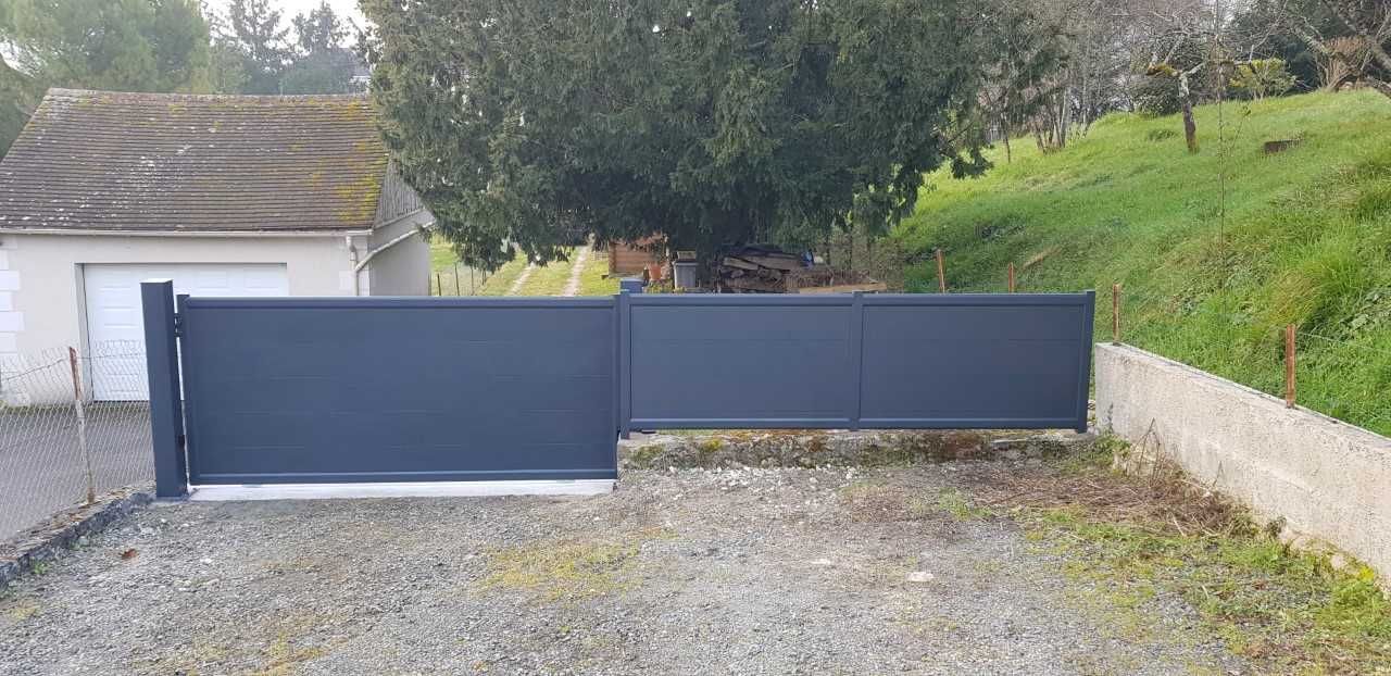 Un portail coulissant bleu enjambe une allée de gravier, à côté d'un garage et d'un talus herbeux.
