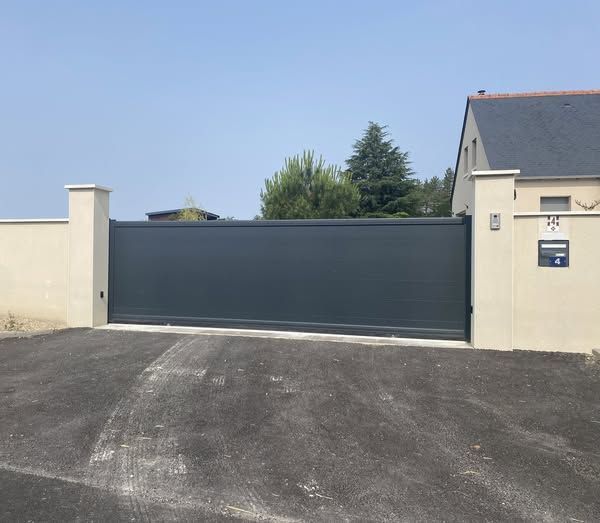 Portail d'allée gris foncé fermé entre des murs couleur crème et une maison, au bord d'une route pavée sous un ciel dégagé