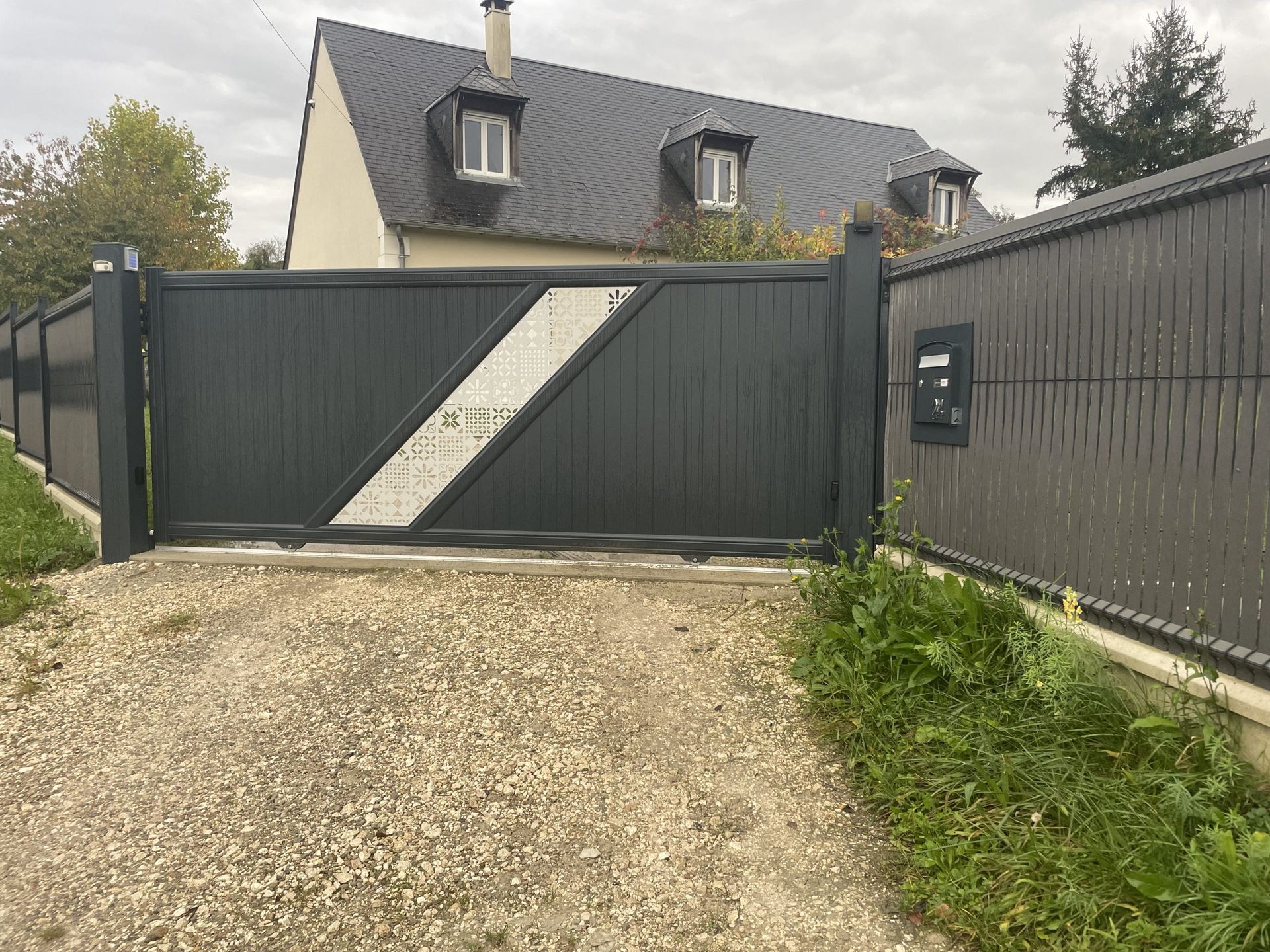 Portail coulissant vert foncé avec une bande blanche diagonale devant une maison et une allée en gravier.