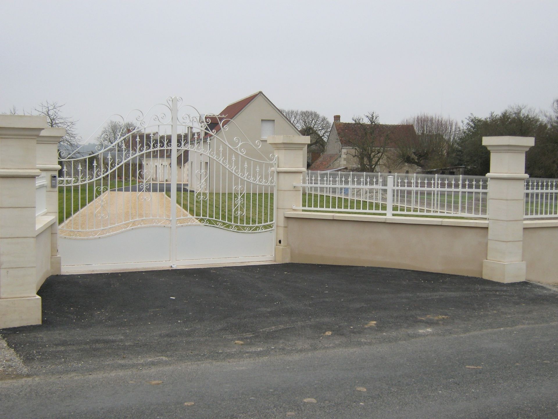 Portail en métal blanc à l'entrée d'une allée carrossable avec des piliers en pierre et une maison en arrière-plan