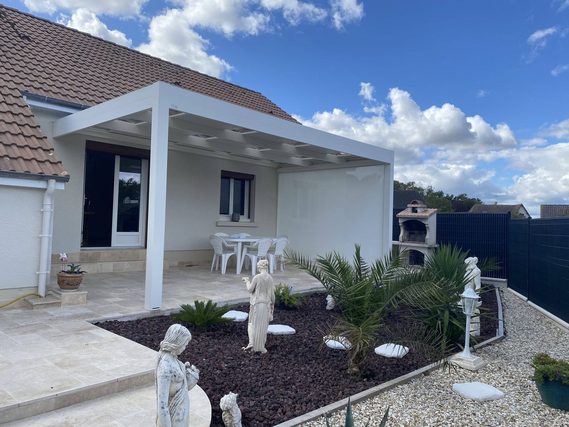 Patio d'une maison blanche avec chaises, plantes en pot et toit de tuiles sous un ciel bleu nuageux