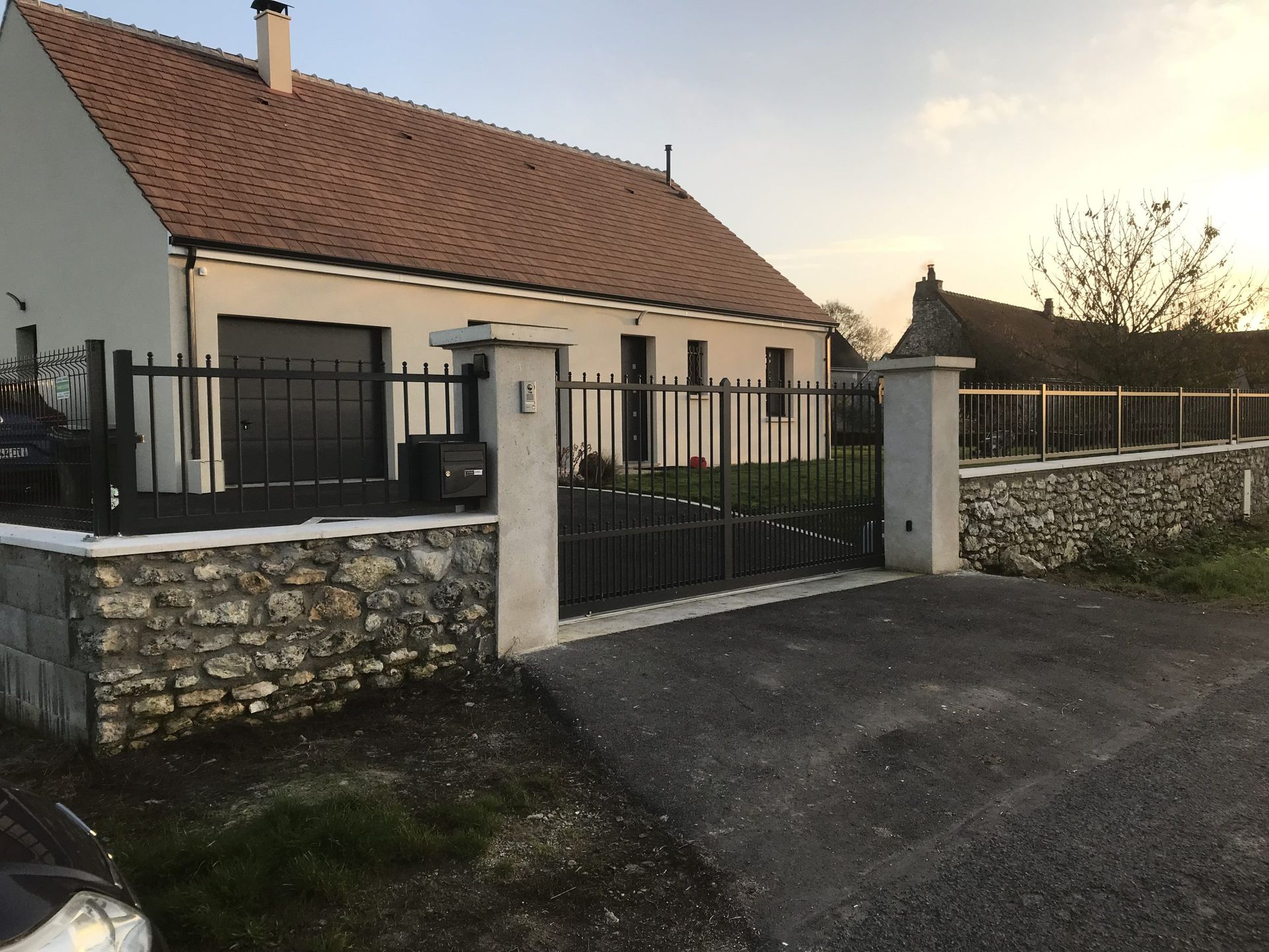 Maison de plain-pied avec allée privée fermée par un portail, mur en pierre et clôture noire au coucher du soleil