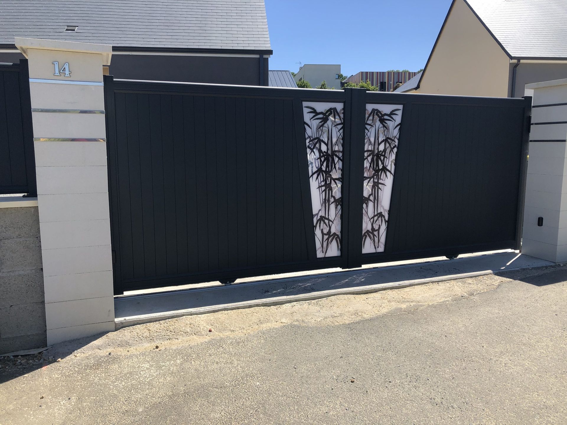 Portail d'allée noir avec deux panneaux décoratifs blancs et noirs entre des piliers en pierre.