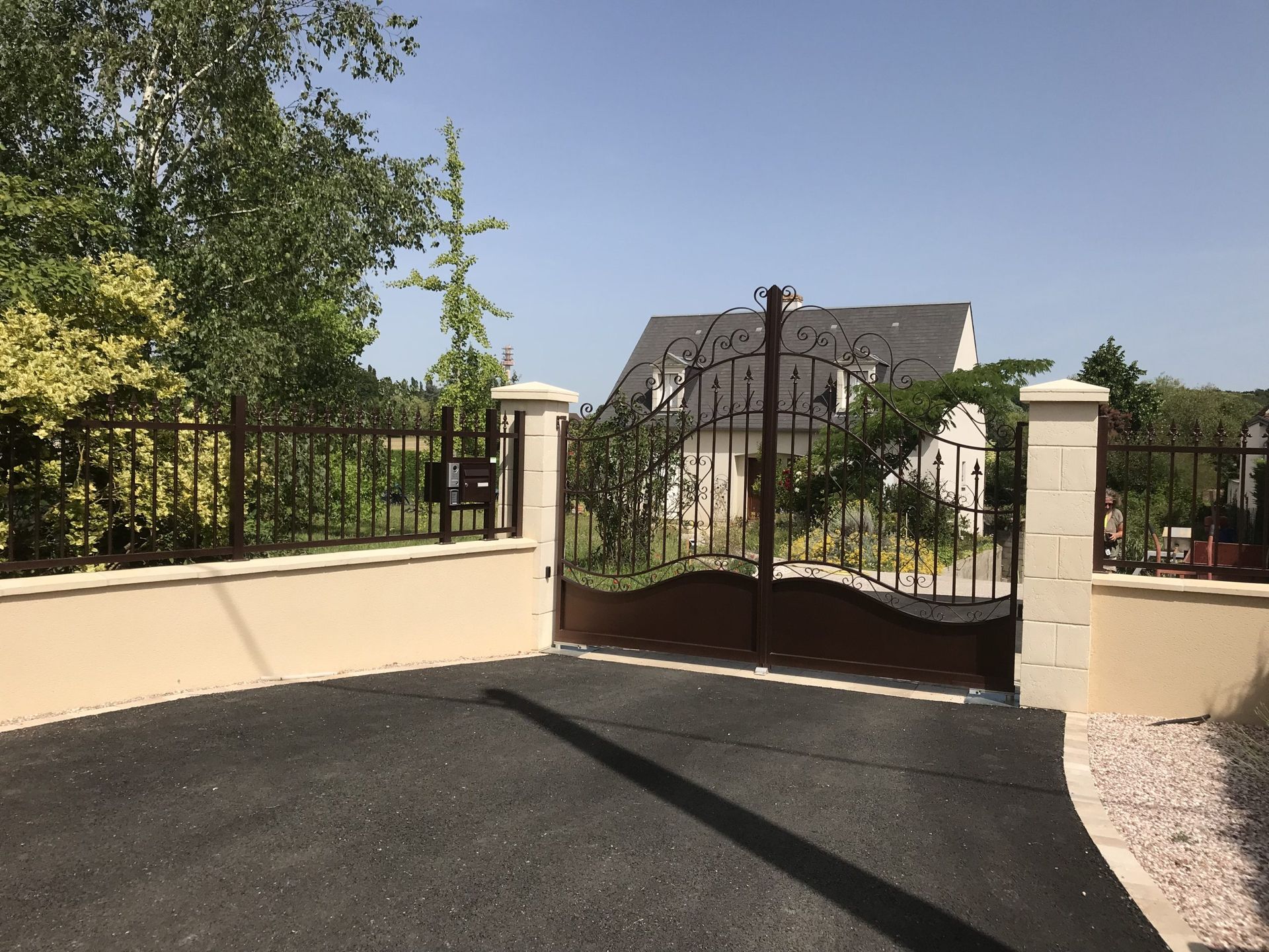 Un portail en fer forgé dans des murs couleur crème s'ouvre sur une allée, avec une maison et des arbres au-delà, sous un ciel dégagé.