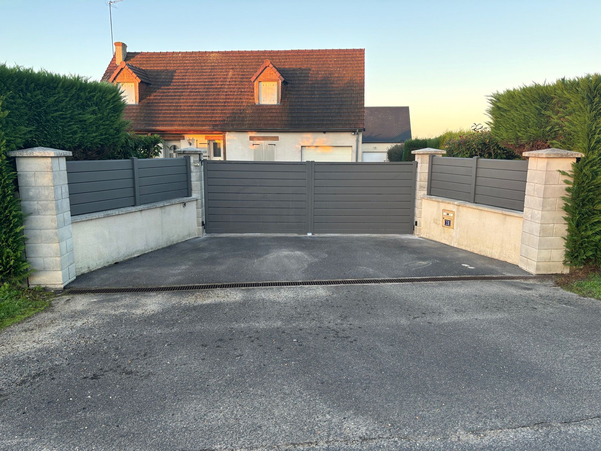 Portail d'allée gris entre des piliers en béton devant une maison au toit de tuiles rouges au crépuscule