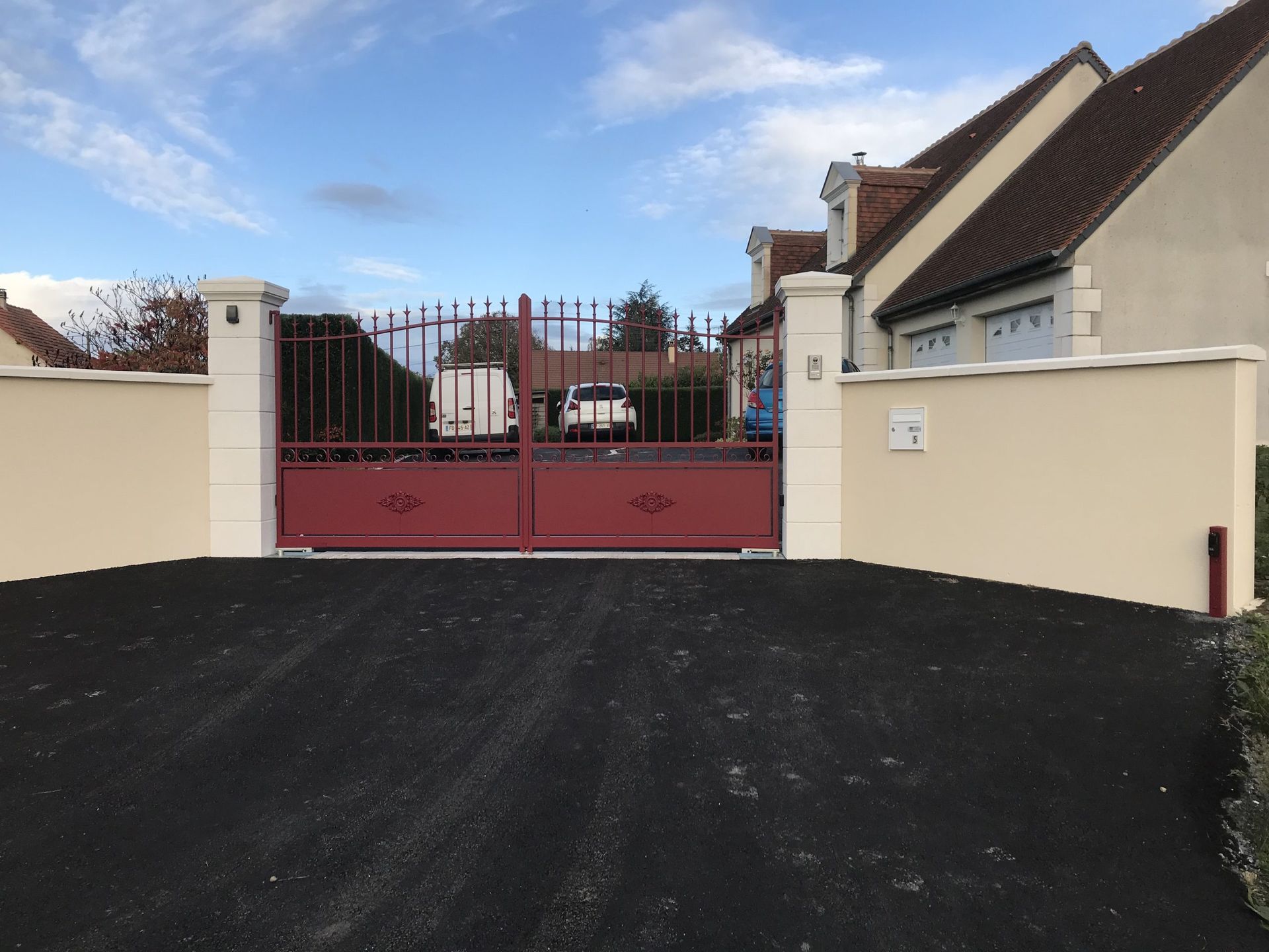 Portail rouge et murs couleur crème au bout d'une allée pavée noire, sous un ciel bleu.