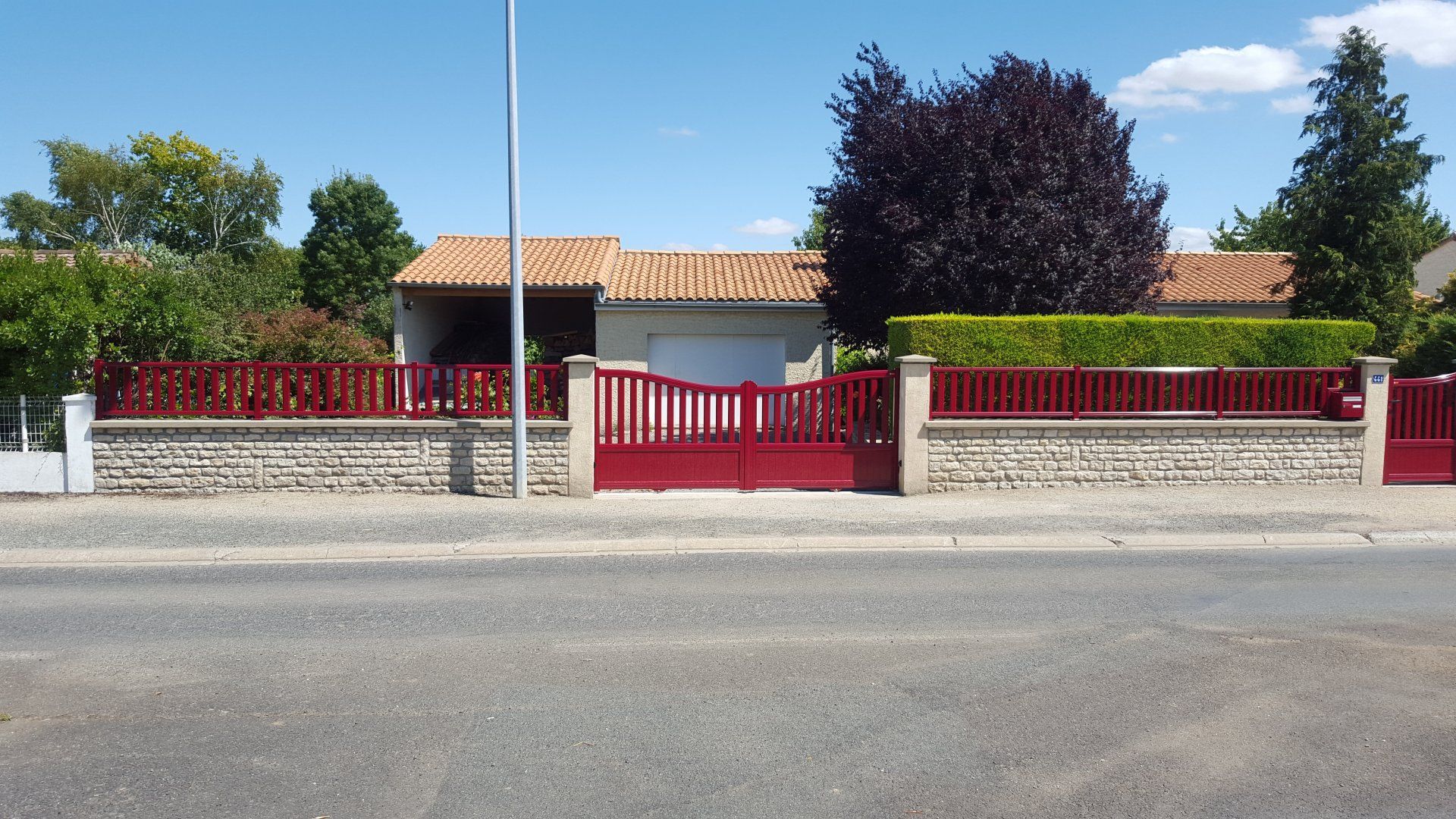 Maison de plain-pied avec portail et clôture rouges, piliers en pierre et arbres en bord de route sous un ciel bleu