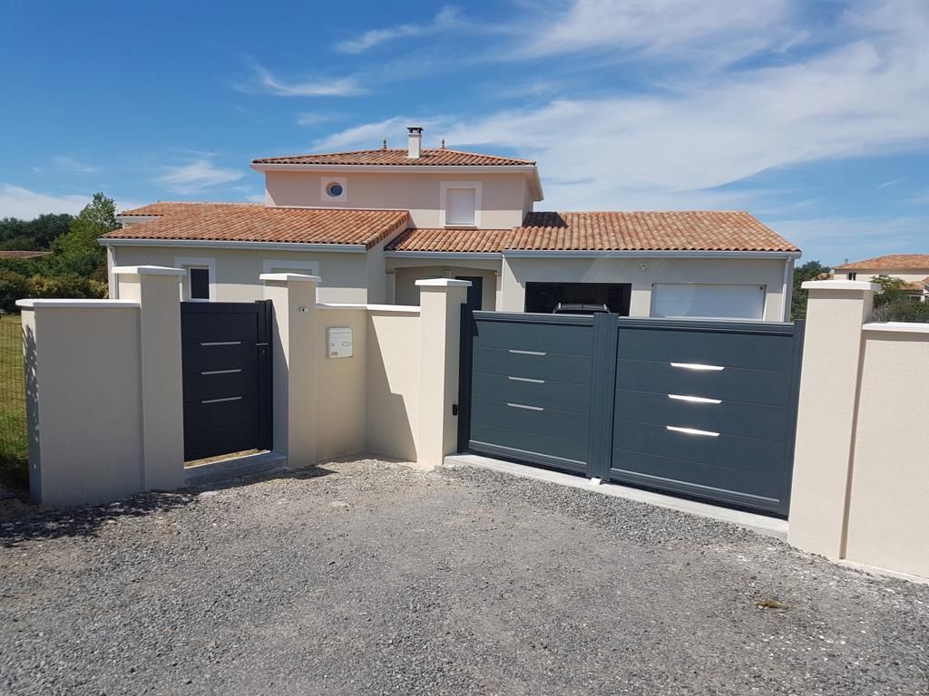 Maison moderne aux murs blancs et au portail gris, donnant sur une allée de gravier sous un ciel bleu.