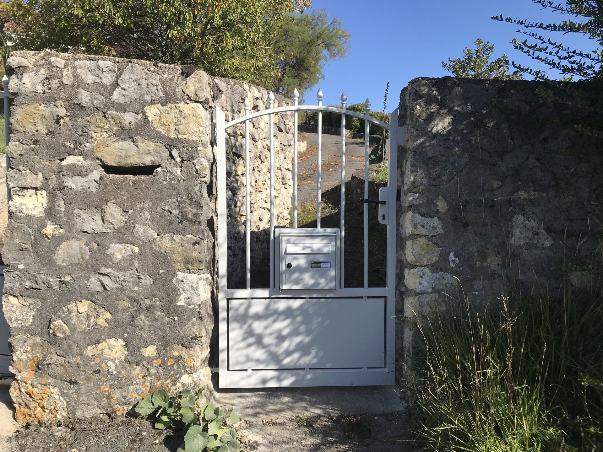 Mur en pierre avec un portail en métal blanc à l'entrée d'un jardin ensoleillé