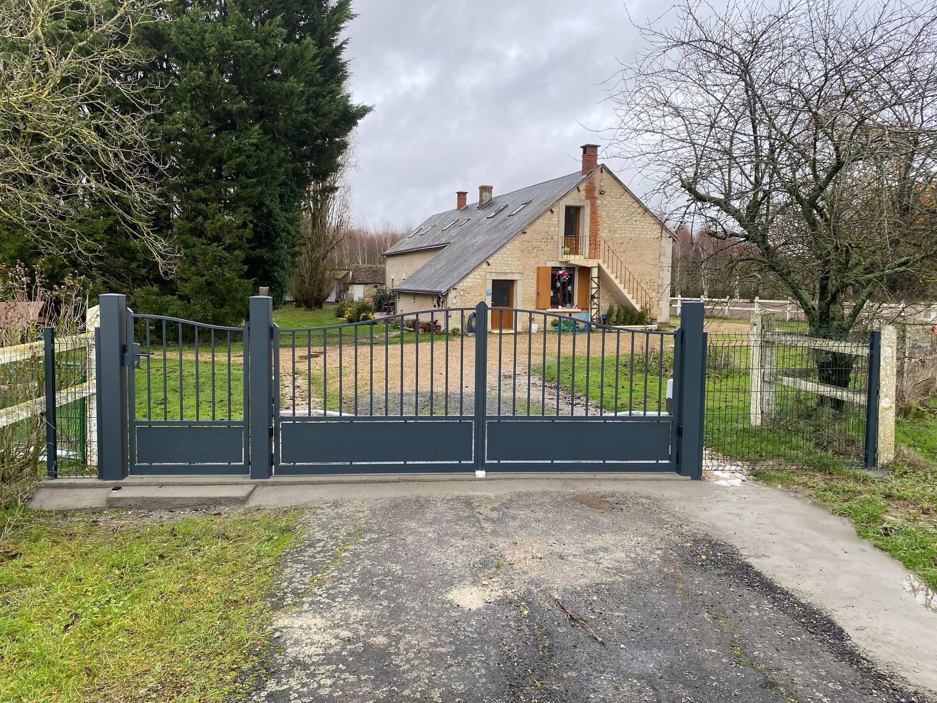 Portail d'allée s'ouvrant sur une maison en pierre avec pelouse et arbres par temps nuageux