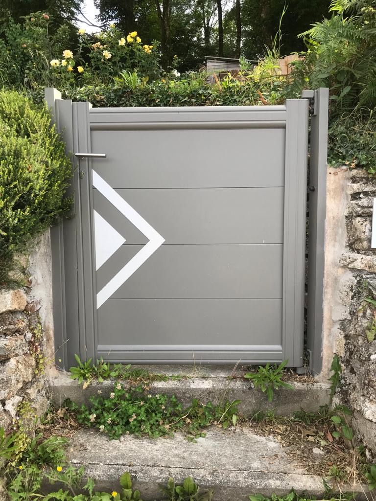 Portillon piétonnier gris orné d'un chevron blanc, encastré dans un mur de pierre au milieu de la verdure.