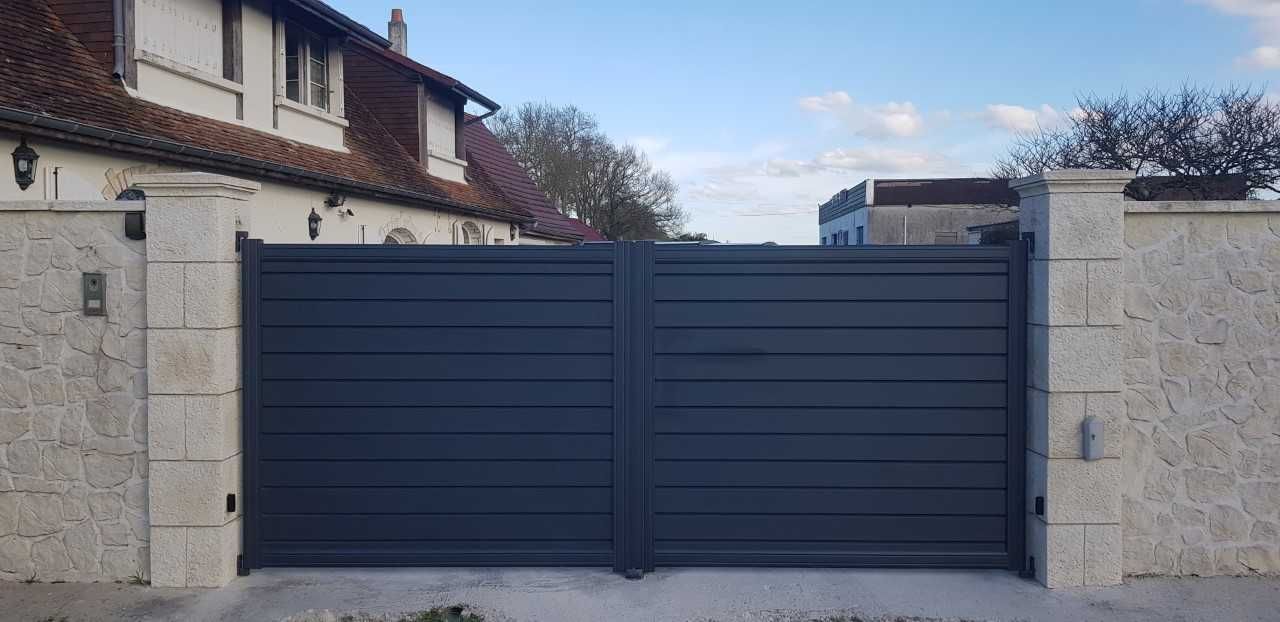 Portail d'allée en métal bleu foncé entre des piliers en pierre devant une maison
