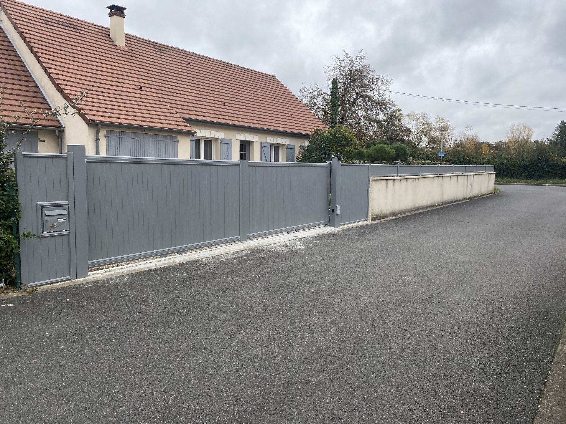 Rue résidentielle tranquille avec une clôture grise, une maison de plain-pied et une route pavée sous un ciel couvert.