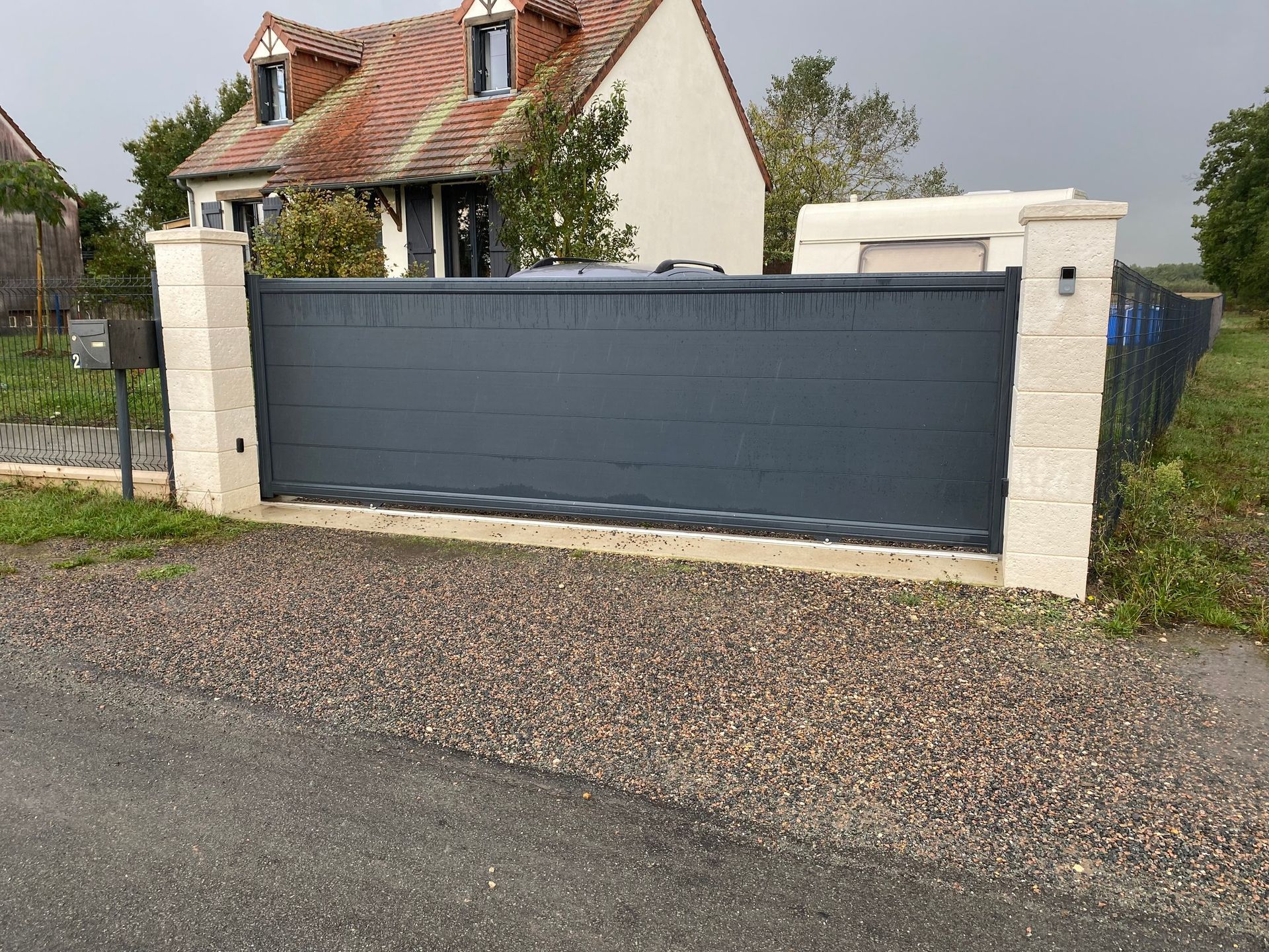 Portail gris et piliers blancs devant une maison au toit de tuiles, le long d'un chemin de gravier.