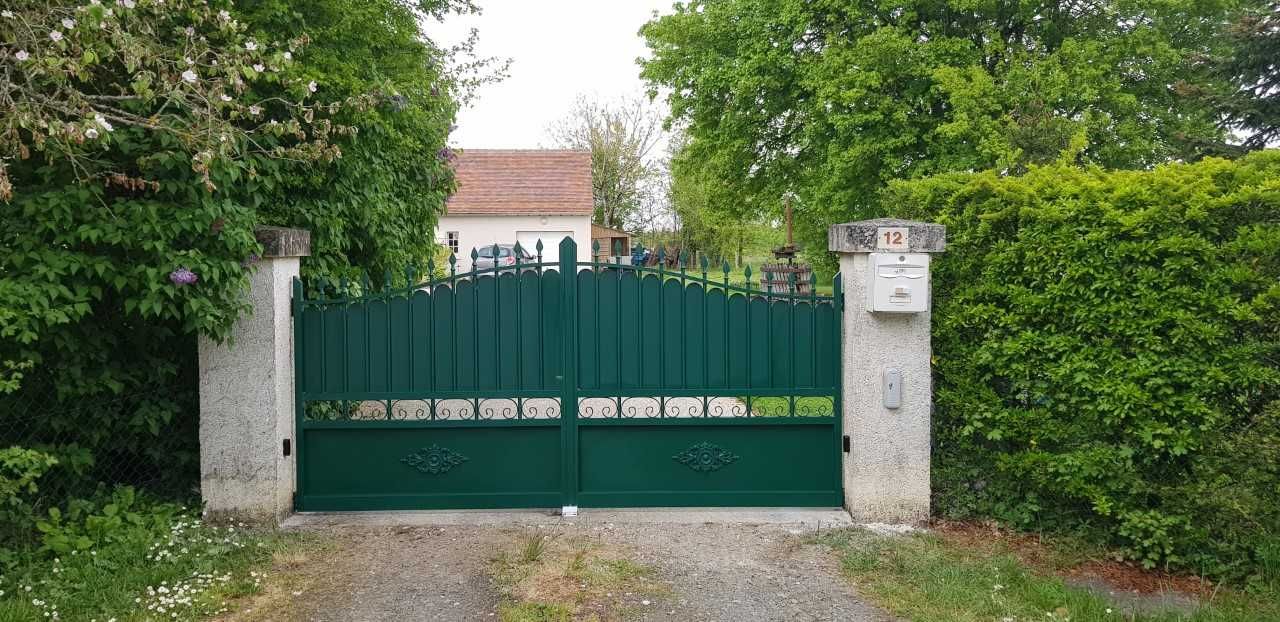 Un portail double vert, encadré de piliers blancs et de haies, bloque l'accès à une maison.