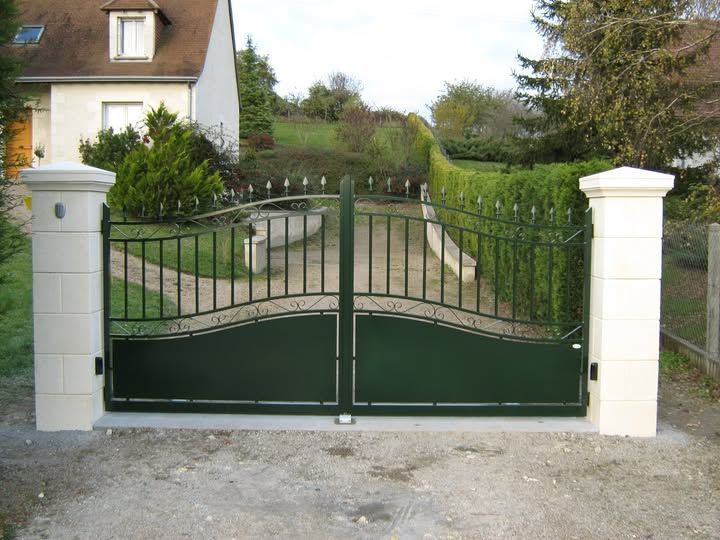 Portail d'allée en métal vert entre des piliers blancs, devant une maison et un jardin.