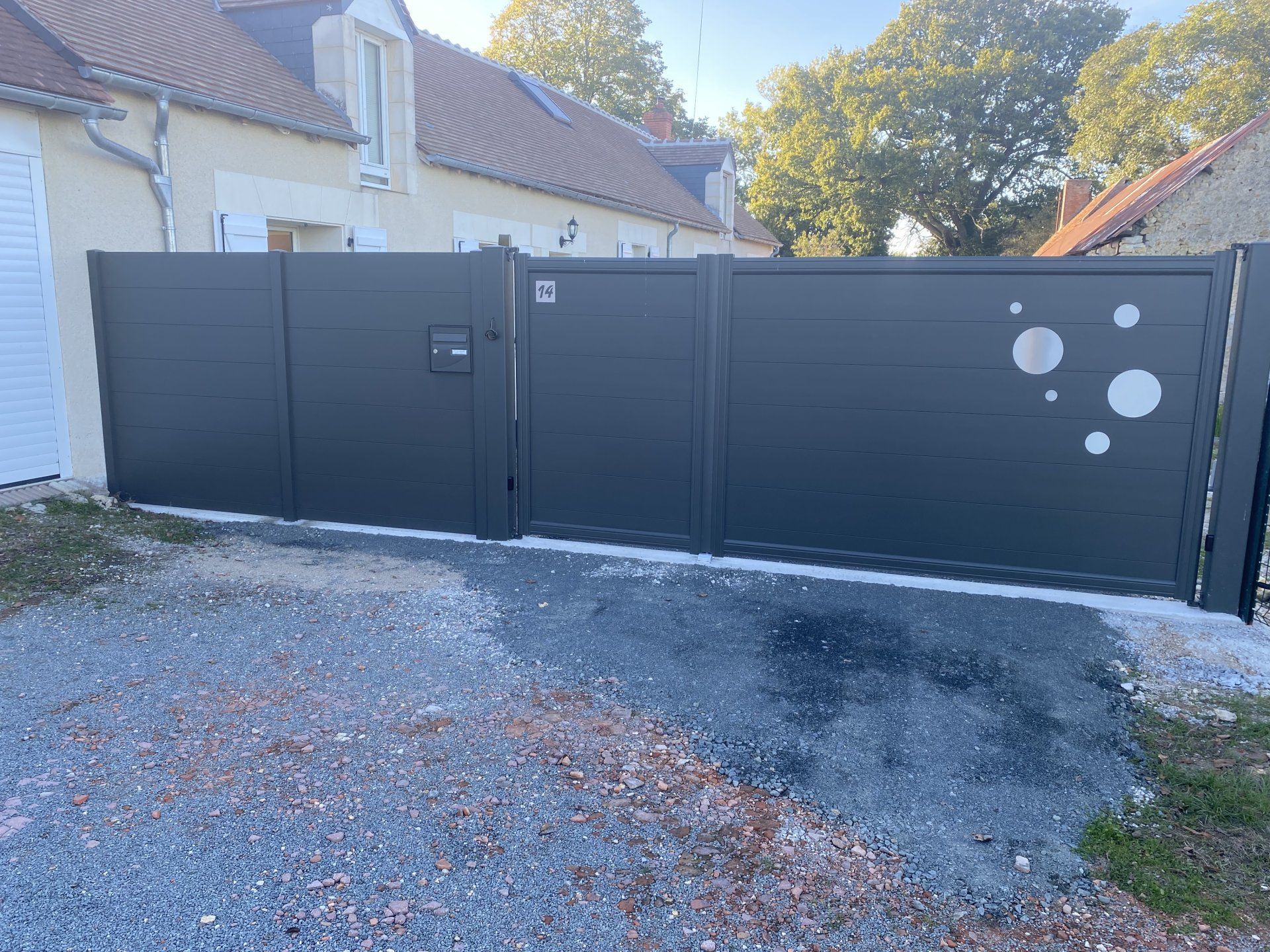 Portail d'allée en métal noir à côté d'une maison, avec un sol en gravier et des arbres en arrière-plan.
