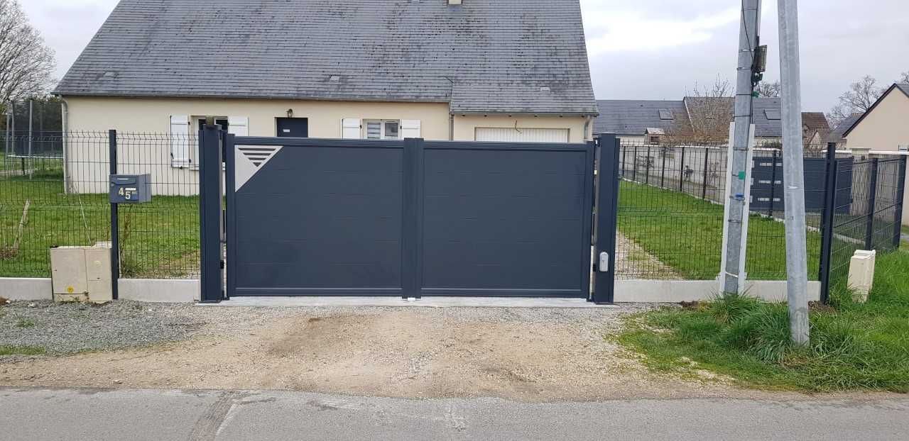 Portail d'allée en métal gris foncé devant une maison bordée de gravier et d'herbe.