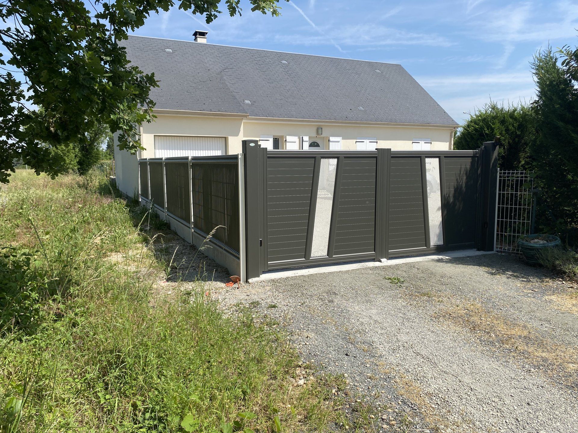 Allée de gravier menant à une clôture et un portail gris foncé devant une maison blanche au toit gris.