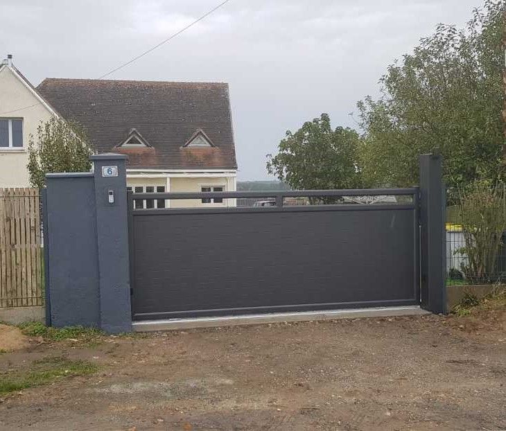 Portail coulissant gris devant une maison au toit en pente et entourée d'arbres.