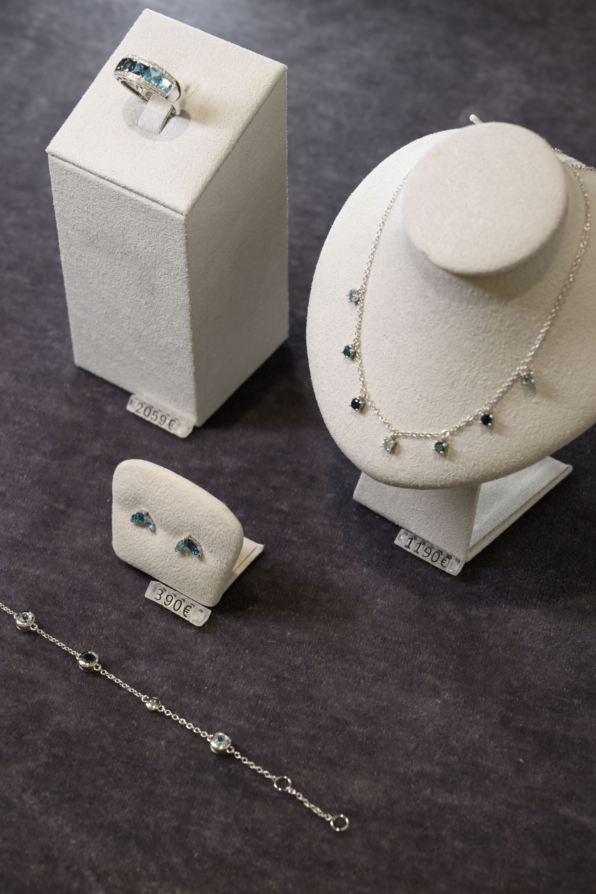 Présentoir à bijoux comprenant une bague, un collier, un bracelet et des boucles d'oreilles ornés de pierres bleu clair sur fond gris.