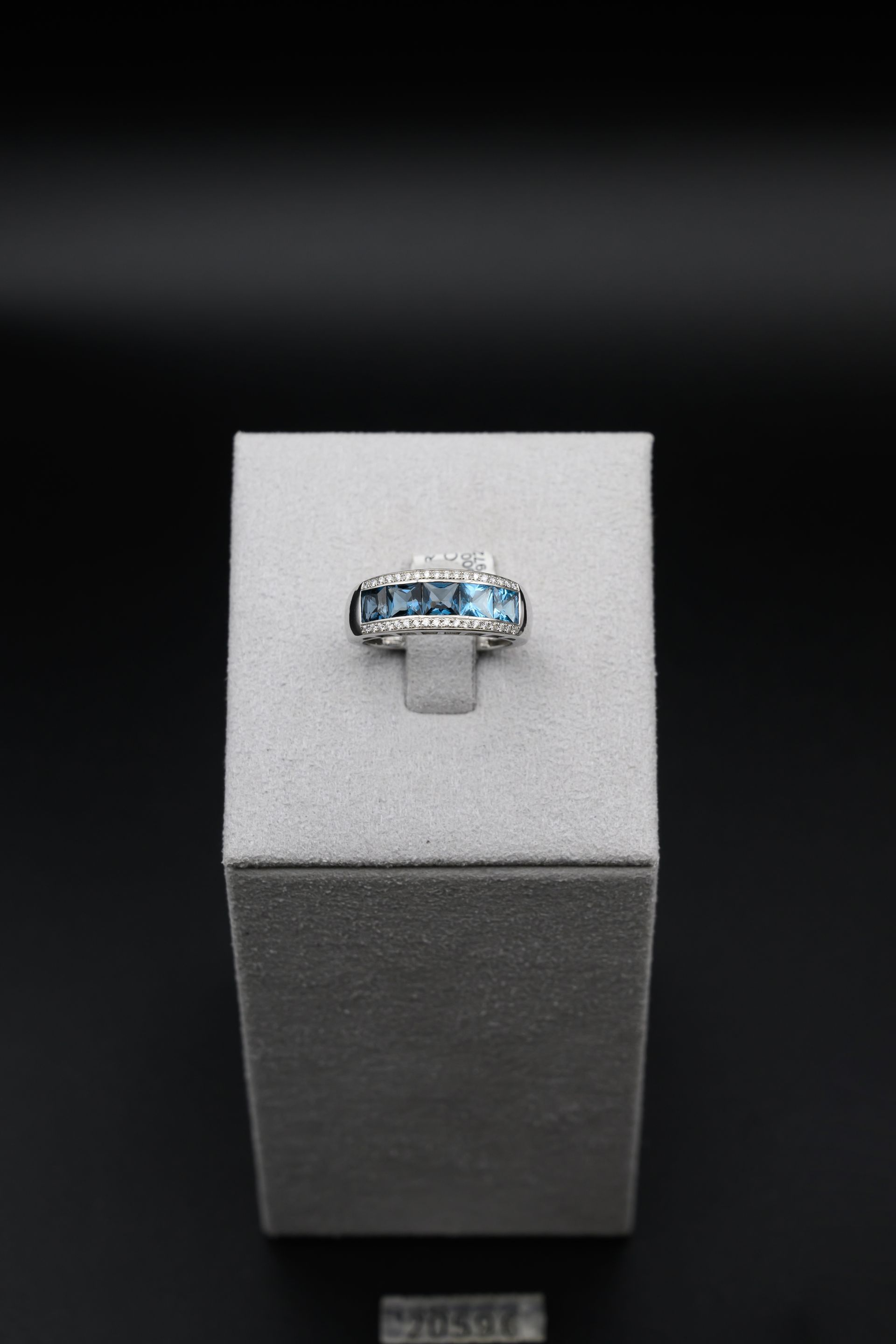 Bague ornée de pierres bleues et de petits diamants, présentée sur un socle gris sur fond noir.
