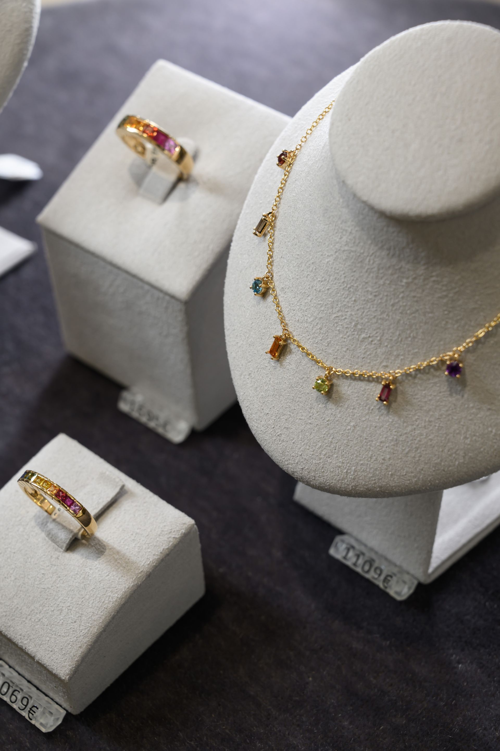 Présentoir à bijoux avec collier et bagues en or ornés de pierres précieuses colorées sur des socles gris.
