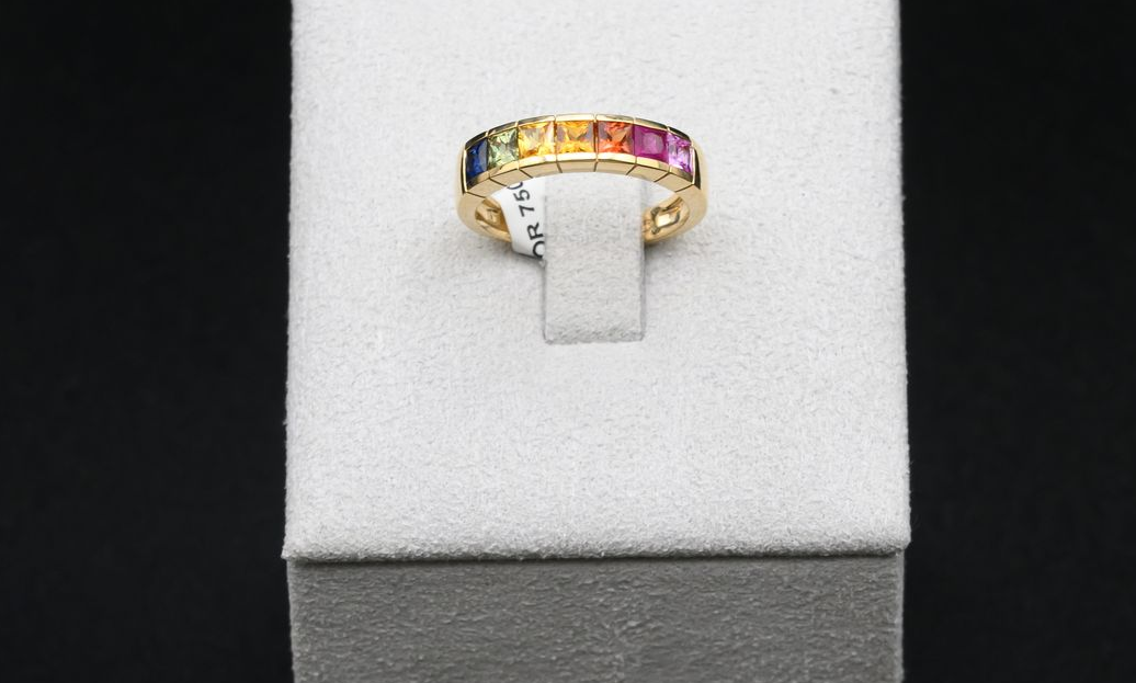 Bague en or ornée d'une rangée de pierres précieuses colorées, sur un support blanc.