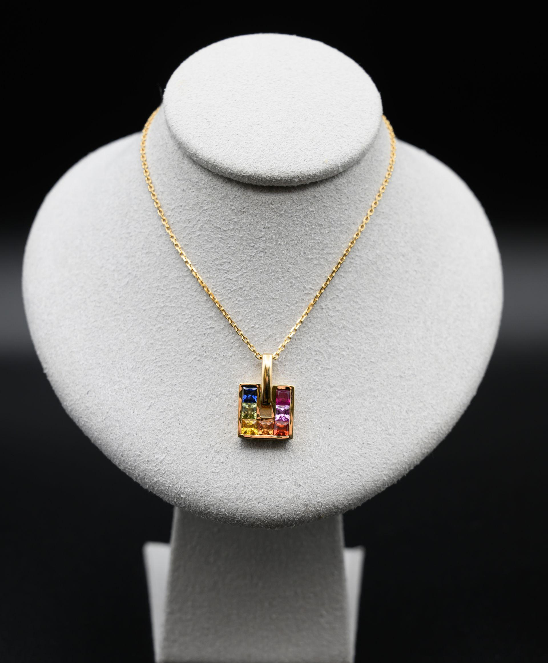 Collier en or avec pendentif carré orné de pierres précieuses colorées, présenté sur un buste blanc sur fond noir.
