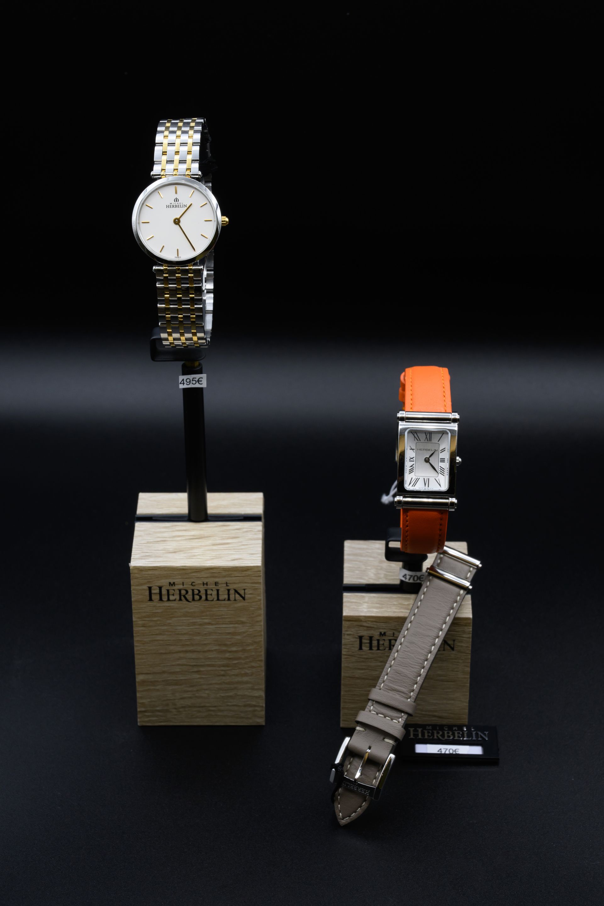 Deux montres sont exposées sur un support devant un fond noir. L'une est ronde et argentée, l'autre carrée avec un bracelet orange.