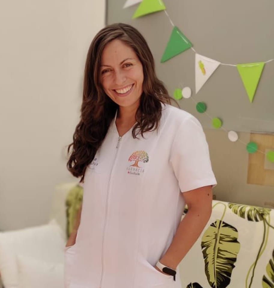 Una mujer con bata blanca sonríe, de pie en un interior con decoraciones coloridas.