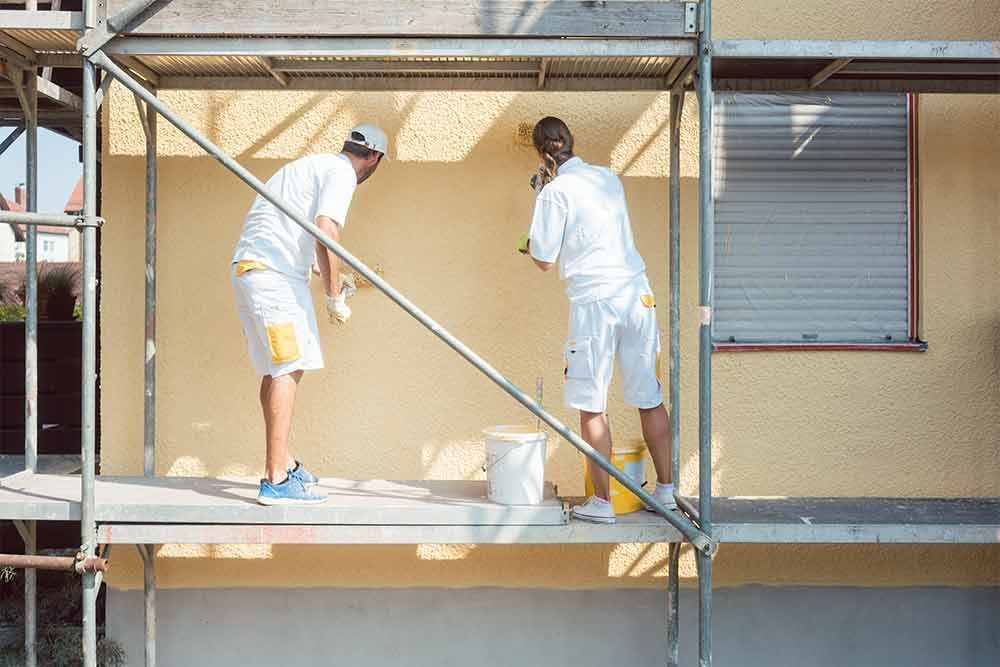 Zwei Männer streichen auf einem Gerüst die Seite eines Hauses.