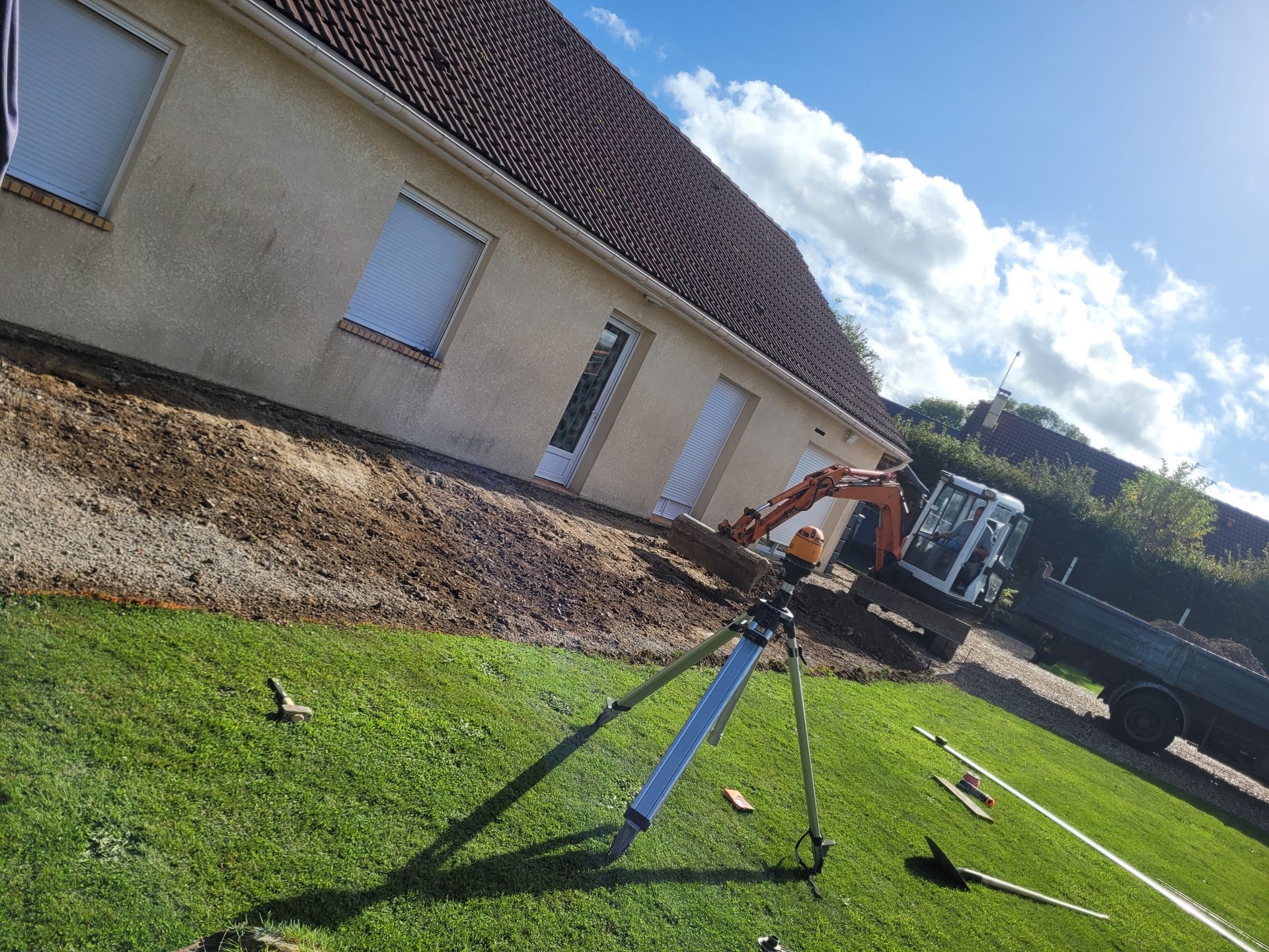 Outil de géomètre posé sur la pelouse face à un terrain préparé pour une terrasse devant un pavillon