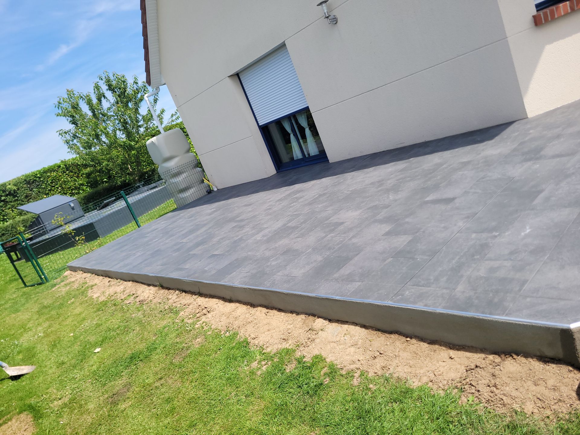 Terrasse dallée couleur béton donnant sur la pelouse d'un jardin et accolée à une maison blanche