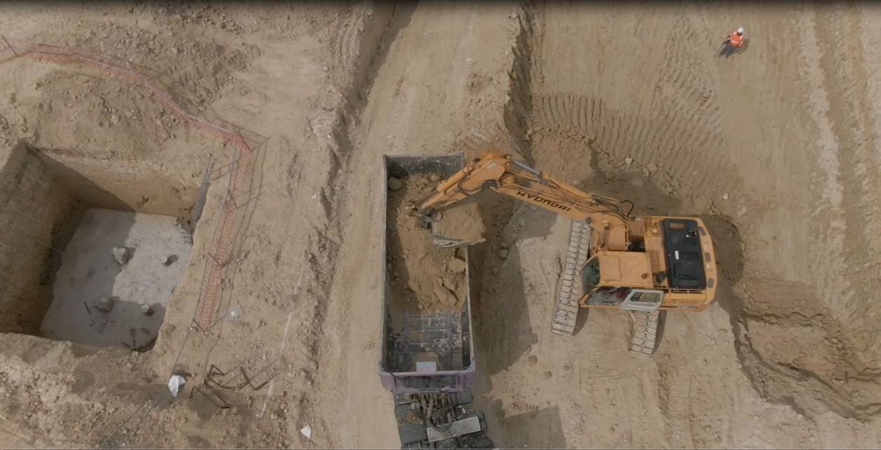 Vista aérea de una excavadora excavando junto a un pozo rectangular profundo en una obra de construcción arenosa.