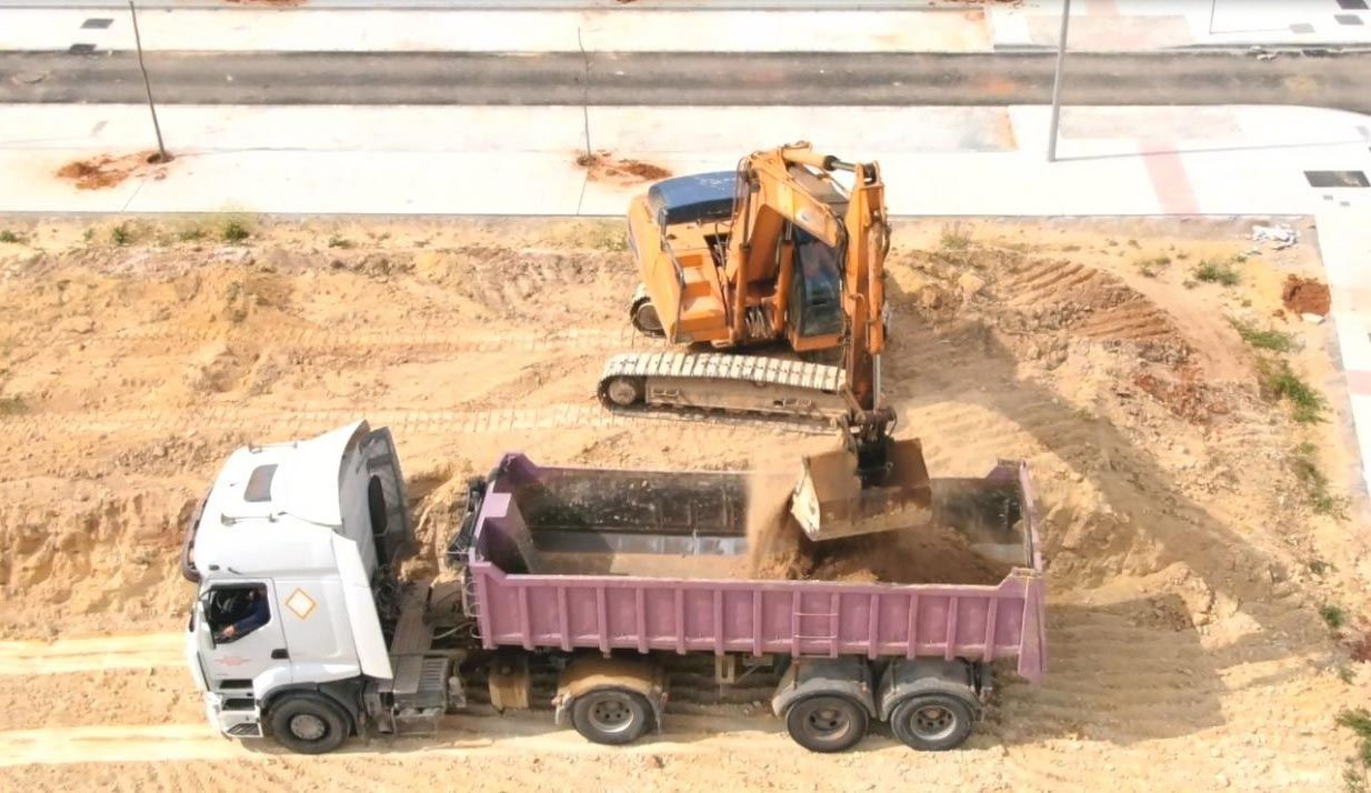 Escena de construcción en miniatura con una excavadora cargando tierra en un camión volquete morado sobre terreno arenoso.