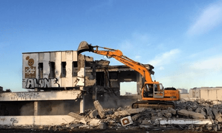 Una excavadora naranja demuele un edificio dañado entre escombros bajo un cielo azul.