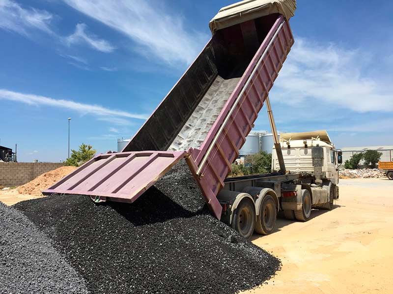 Camión volquete descargando una pila de grava negra en una obra de construcción bajo un cielo azul.