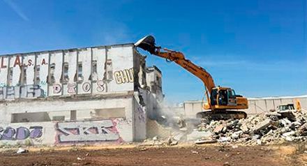 Una excavadora demuele un edificio de hormigón, dejando escombros y polvo bajo un cielo azul despejado.