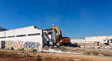 Excavadora de demolición derribando un edificio cubierto de grafitis bajo un cielo azul despejado.
