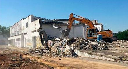Una excavadora demuele un edificio blanco, con escombros apilados delante bajo un cielo azul despejado.